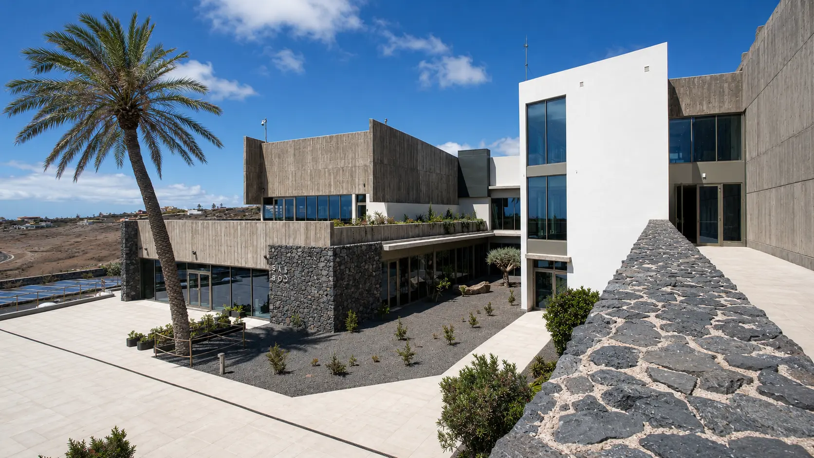 Edificio principal del hotel con fachadas blancas, hormigón, piedra basáltica y una palmera
